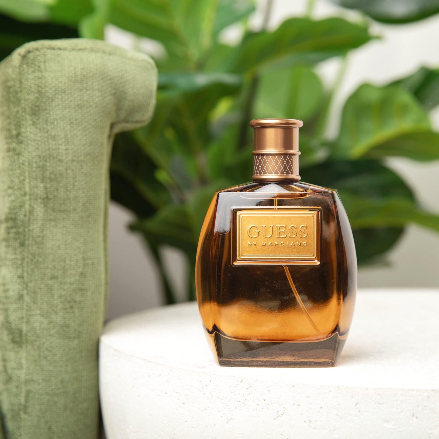 Guess by Marciano Eau De Toilette amber-colored perfume bottle placed on a white textured surface with green plants and a velvet chair in the background