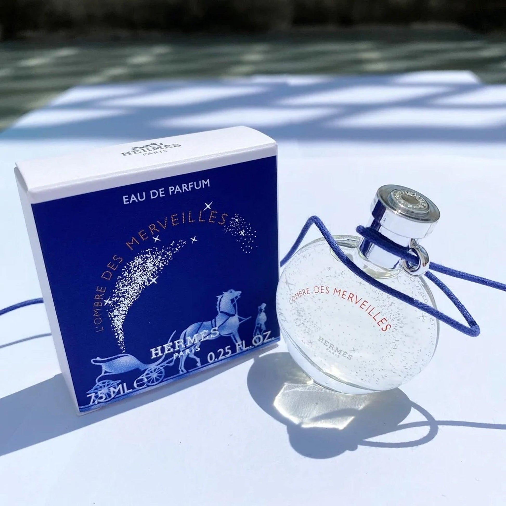 Hermès L’Ombre Des Merveilles Eau De Parfum mini bottle and box placed on a white surface under natural sunlight with blue shadows.