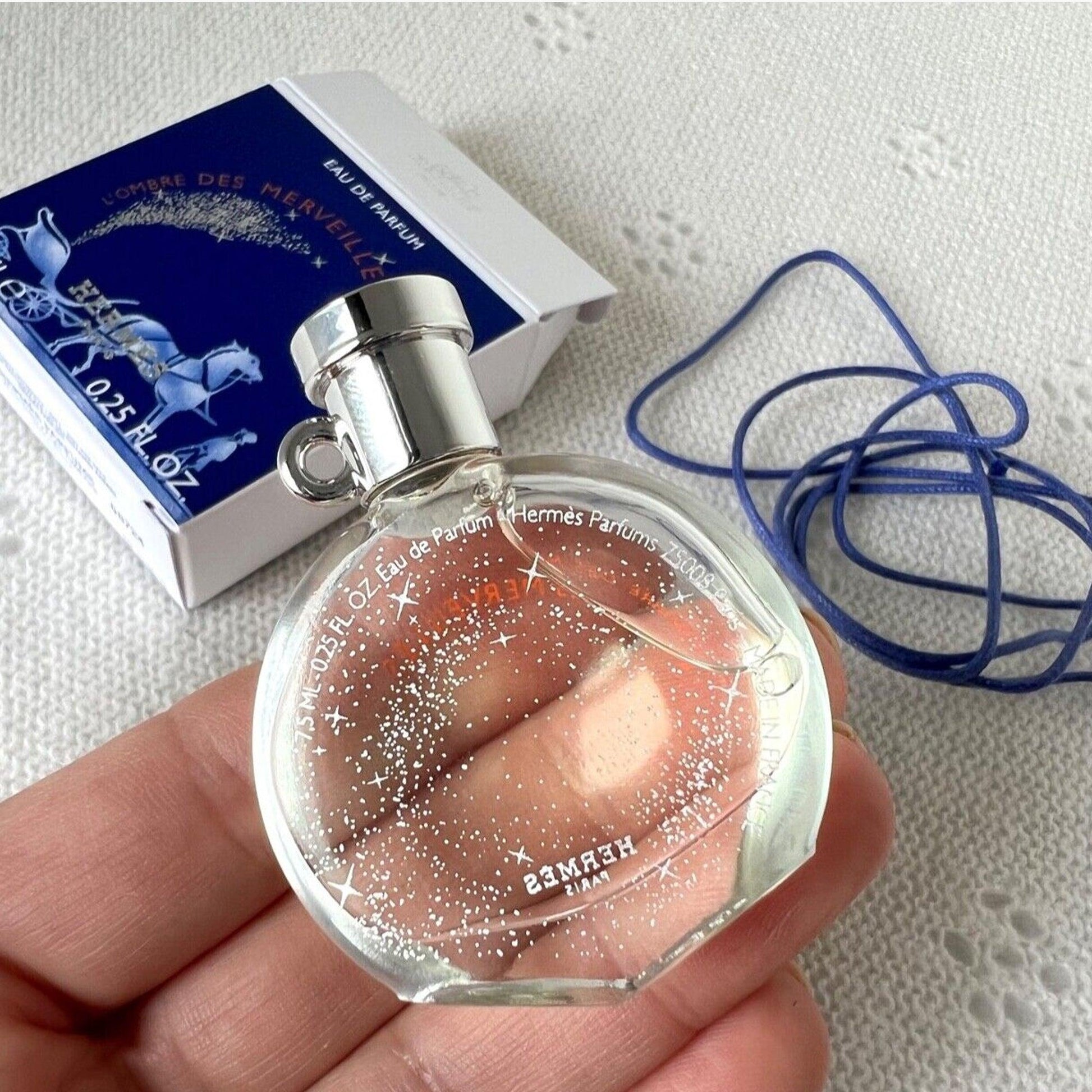 Hand holding a Hermès L’Ombre Des Merveilles Eau De Parfum mini bottle with its box and blue cord placed on a white textured surface.