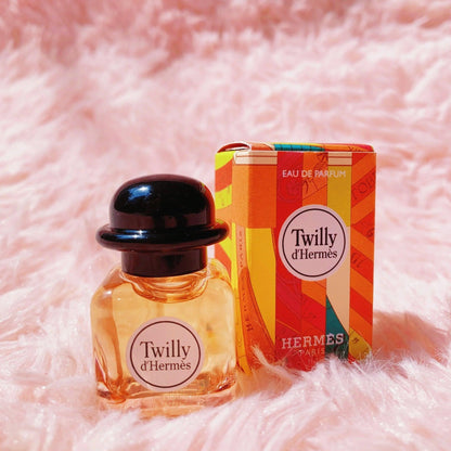 Hermès Twilly d’Hermès Eau De Parfum mini bottle with black cap placed beside its colorful orange patterned box on a soft pink fur background