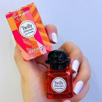 Hand holding Hermès Twilly d’Hermès Eau Poivrée Eau De Parfum mini bottle and matching pink-orange box against a light background.