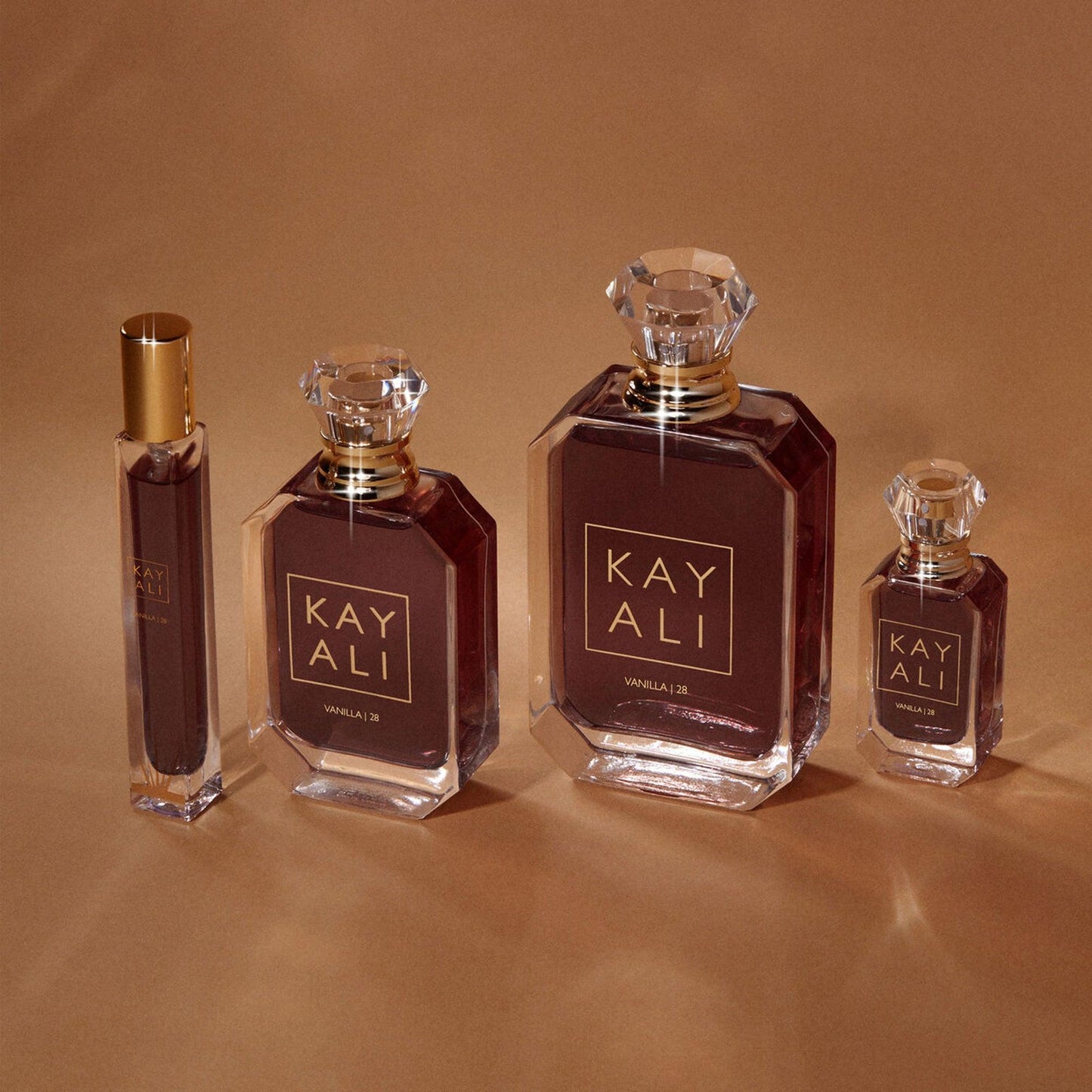 Kayali Vanilla 28 Eau De Parfum bottle collection in different sizes displayed on a warm beige background, showcasing its elegant amber liquid and crystal-cut caps.
