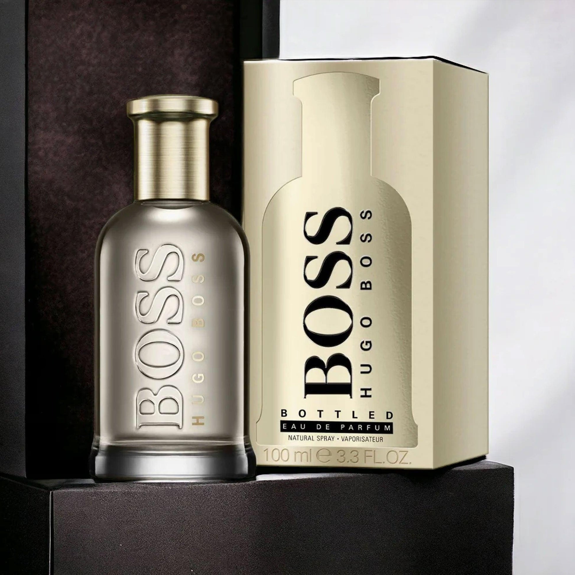 Hugo Boss Bottled Eau De Parfum 100ml for men featuring a sleek metallic bottle placed beside its gold packaging box on a dark background.