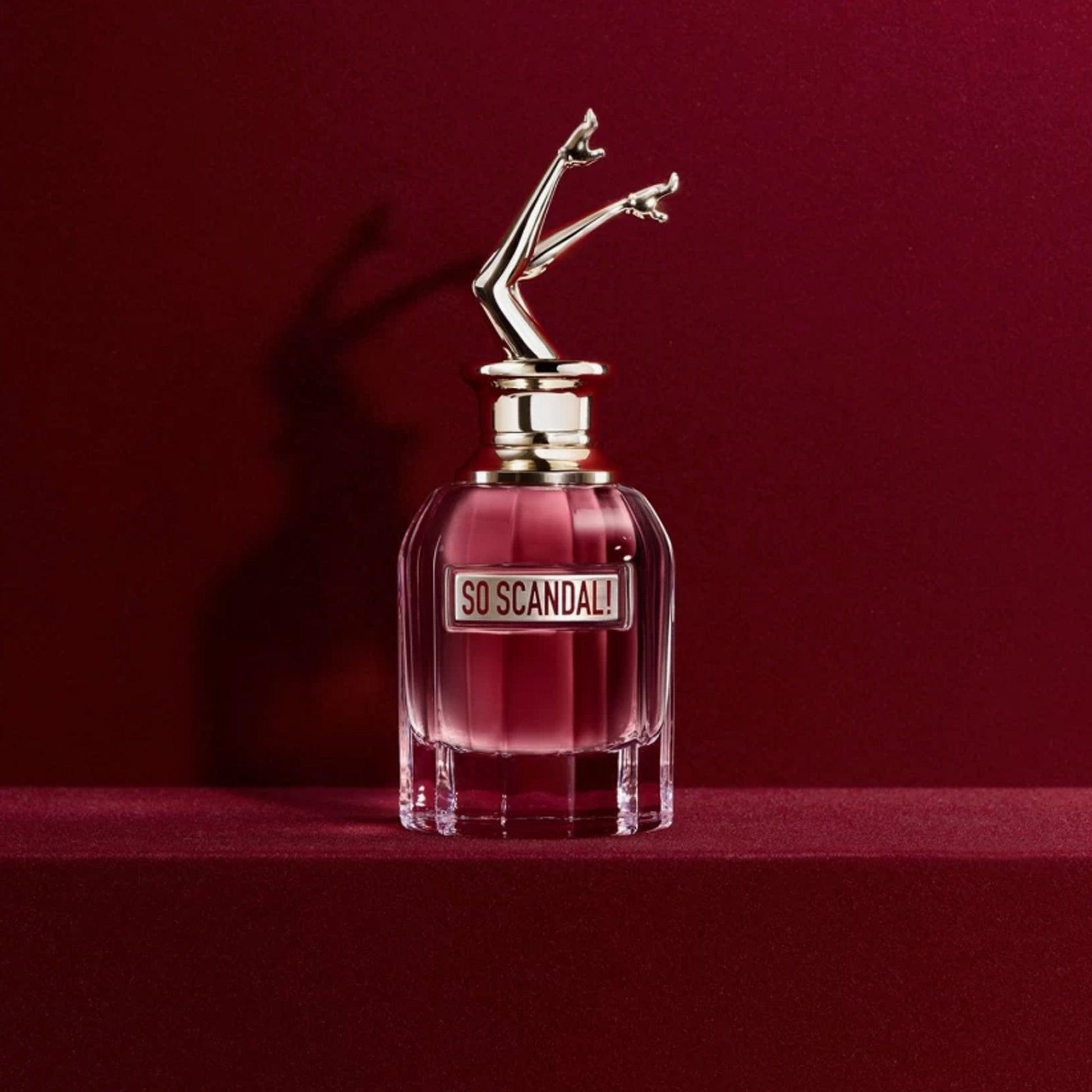 Jean Paul Gaultier So Scandal! Eau De Parfum with a playful leg-shaped cap, placed on a red velvet surface with a deep red background