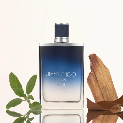 Jimmy Choo Man Blue Eau De Toilette bottle with a blue gradient design placed beside fresh green leaves and a piece of wood on a light reflective surface.