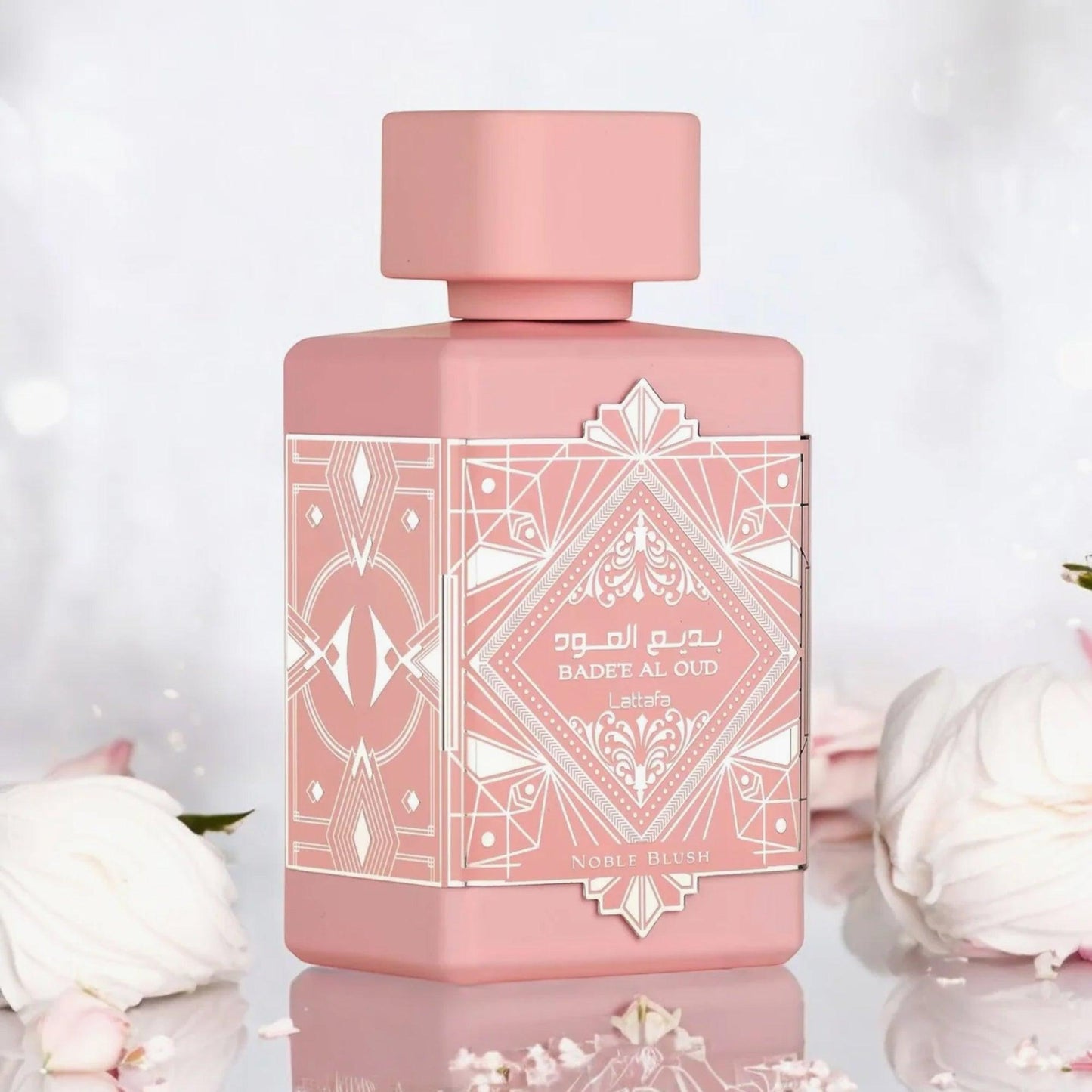 Lattafa Badee Al Oud Noble Blush Eau De Parfum pink bottle with intricate geometric design placed on a reflective surface surrounded by soft white roses and petals