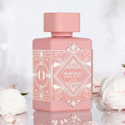Lattafa Badee Al Oud Noble Blush Eau De Parfum pink bottle with intricate geometric design placed on a reflective surface surrounded by soft white roses and petals