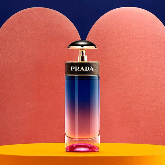 Prada Candy Night Eau De Parfum bottle with gradient pink-to-blue design placed on an orange surface against a pink and navy abstract background