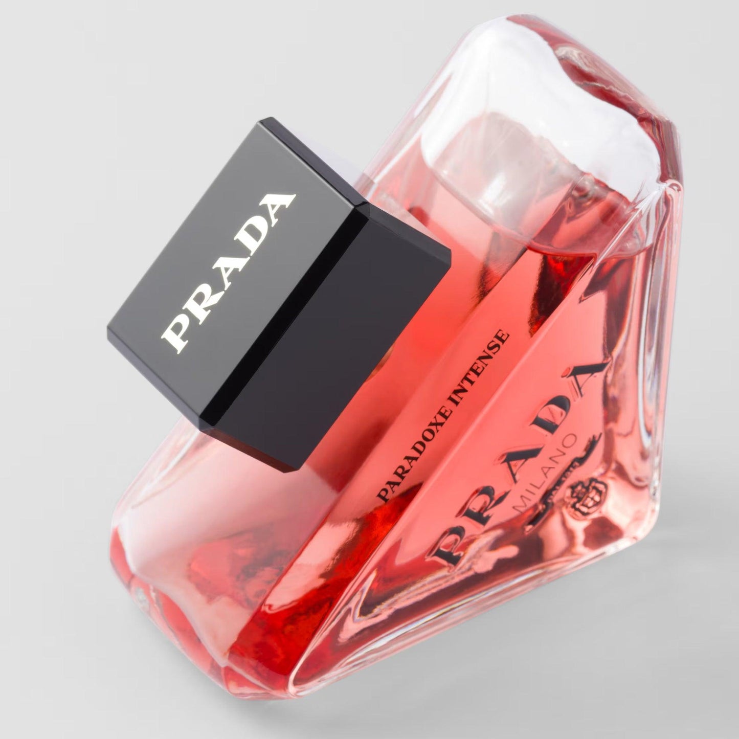 Prada Paradoxe Intense perfume bottle in a bold triangular design with pink-red liquid and a black cap, placed on a light grey background