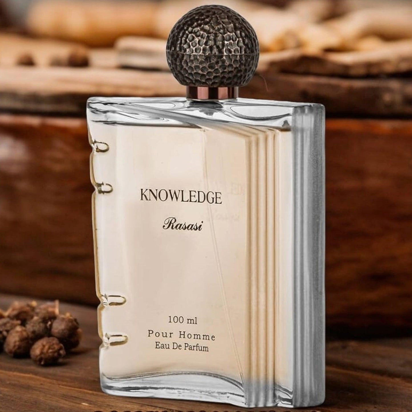 Rasasi Knowledge Pour Homme Eau De Parfum 100ml bottle with a unique book-shaped design, displayed on a wooden surface surrounded by warm spices for a rich masculine aesthetic