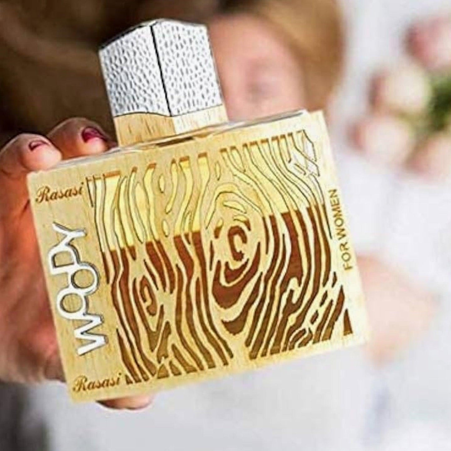 Rasasi Woody for Women Eau De Parfum bottle with a wood-grain inspired golden design held in hand against a soft blurred background.