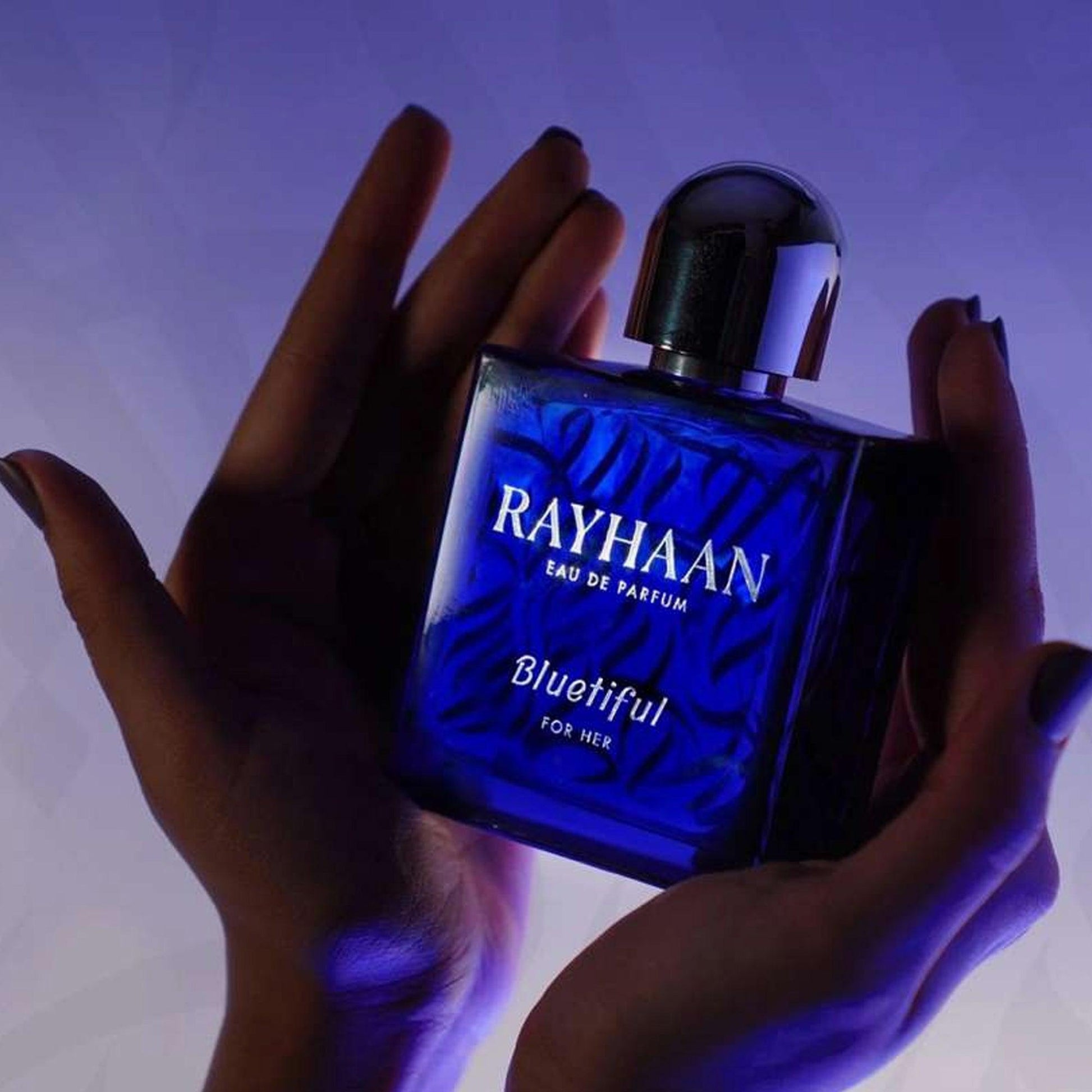 Rayhaan Bluetiful For Her Eau De Parfum blue glass bottle held gently between hands under soft purple lighting