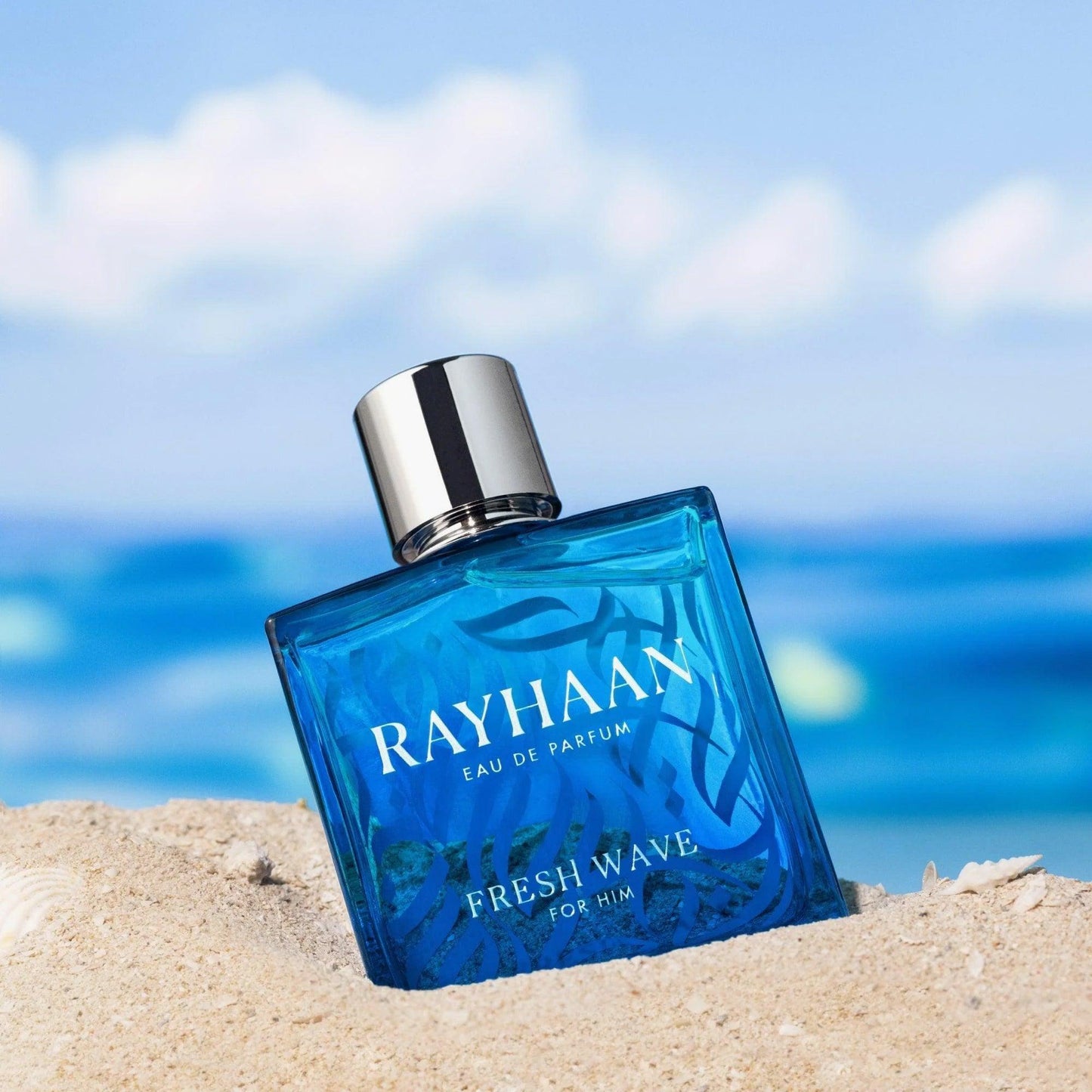 Rayhaan Fresh Wave For Him Eau De Parfum blue glass bottle with silver cap placed on beach sand with the ocean and sky in the background