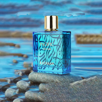 Rayhaan Pacific Pour Homme Eau De Parfum blue patterned glass bottle with gold cap placed on wet stones beside calm ocean water background