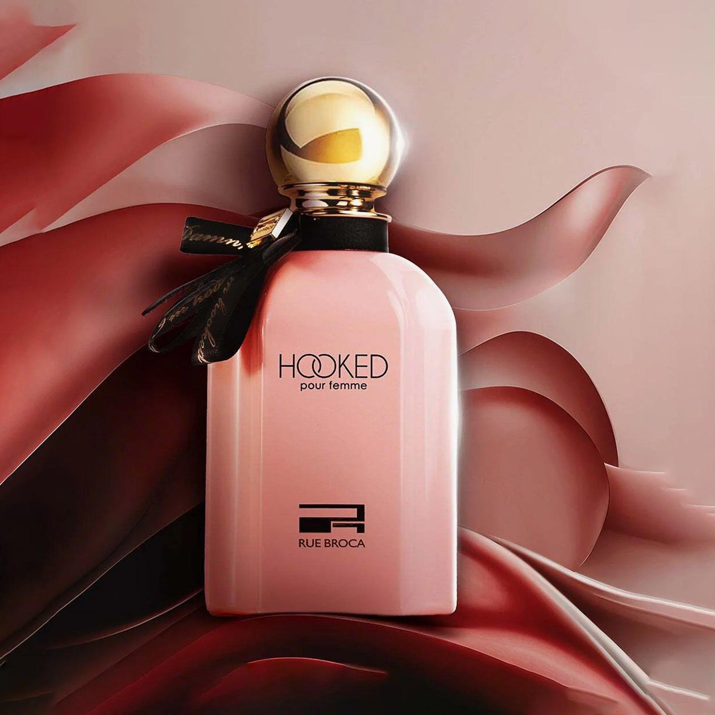 Rue Broca Hooked Pour Femme Eau De Parfum pink bottle with gold cap and black ribbon placed against a flowing satin fabric background in red and beige tones