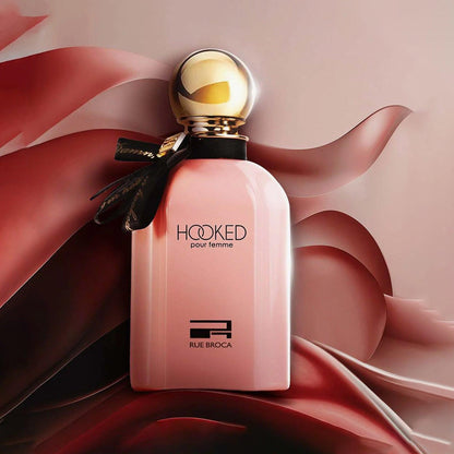 Rue Broca Hooked Pour Femme Eau De Parfum pink bottle with gold cap and black ribbon placed against a flowing satin fabric background in red and beige tones