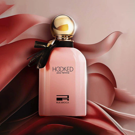 Rue Broca Hooked Pour Femme Eau De Parfum pink bottle with gold cap and black ribbon placed against a flowing satin fabric background in red and beige tones