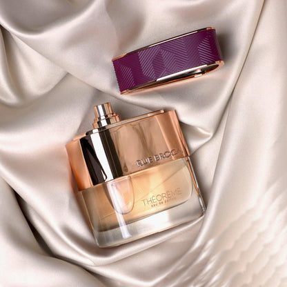Rue Broca Theoreme Eau De Parfum bottle with gold and transparent design placed on silky beige fabric beside its purple and gold cap