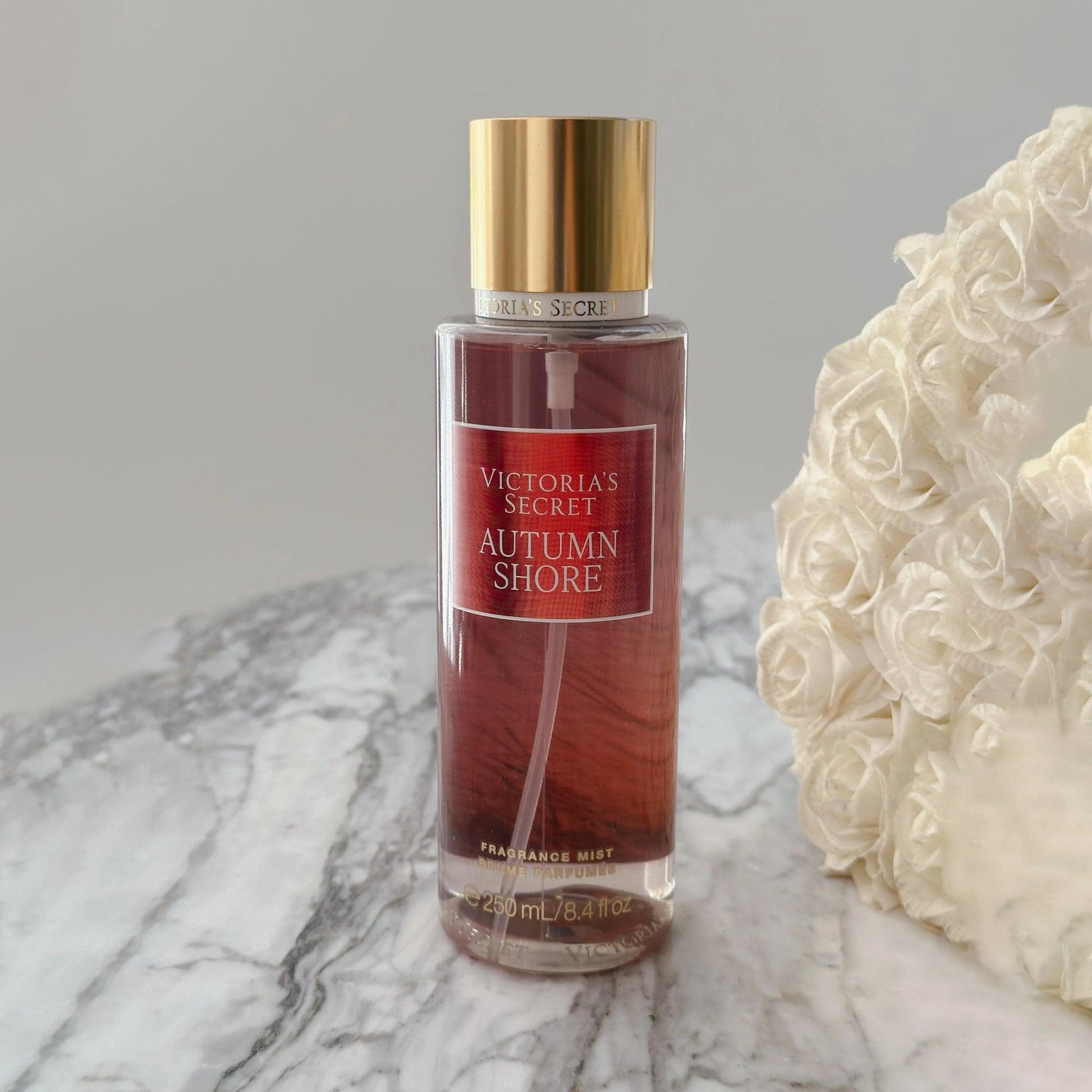 Victoria’s Secret Autumn Shore fragrance mist 250ml bottle with golden cap placed on marble surface beside white rose bouquet