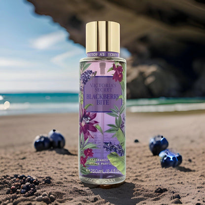Victoria’s Secret Blackberry Bite fragrance mist 250ml bottle placed on sandy beach surrounded by blackberries with ocean waves and rocky cliffs in the background