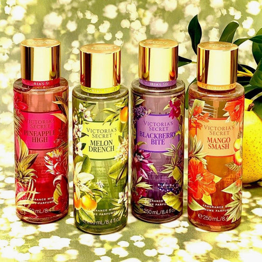 Victoria’s Secret body mist collection including Pineapple High, Melon Drench, Blackberry Bite, and Mango Smash fragrance bottles arranged on a reflective surface with floral background