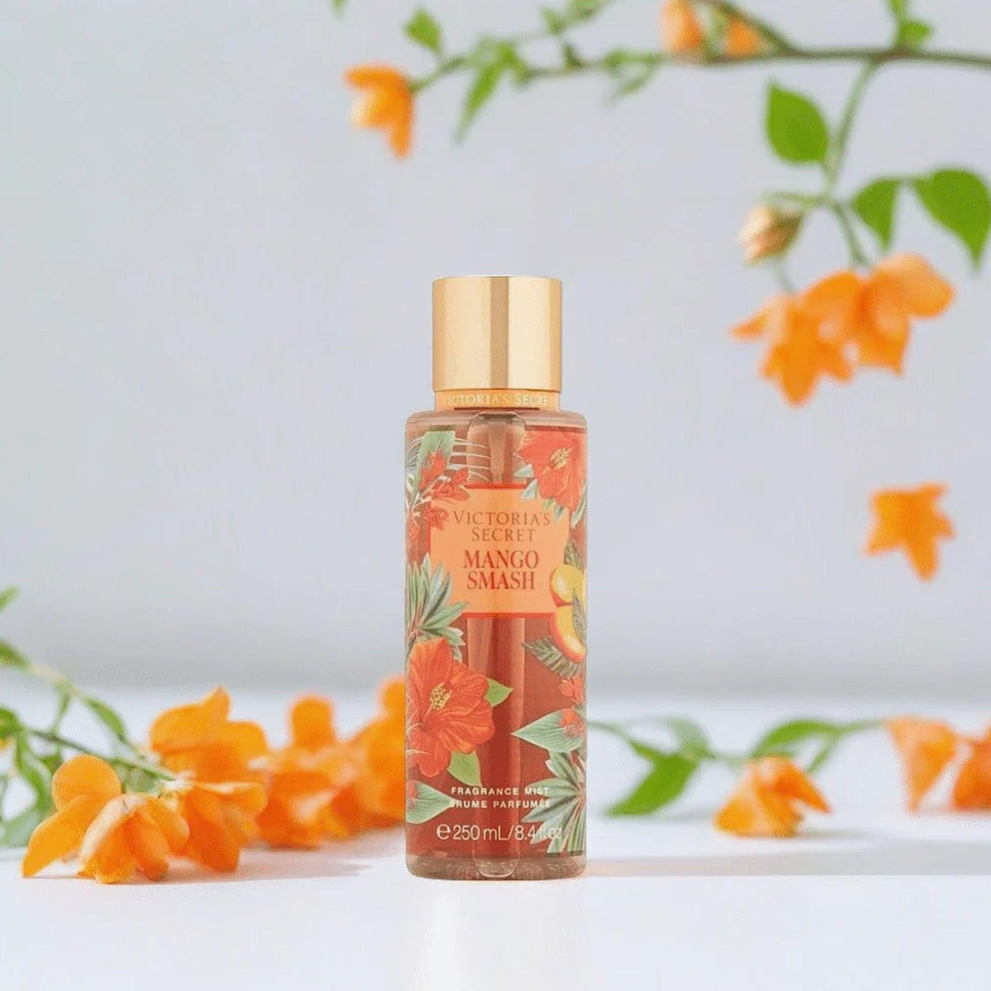 Victorias Secret Mango Smash fragrance mist 250ml bottle with tropical floral design placed on a white surface surrounded by orange flowers and green leaves
