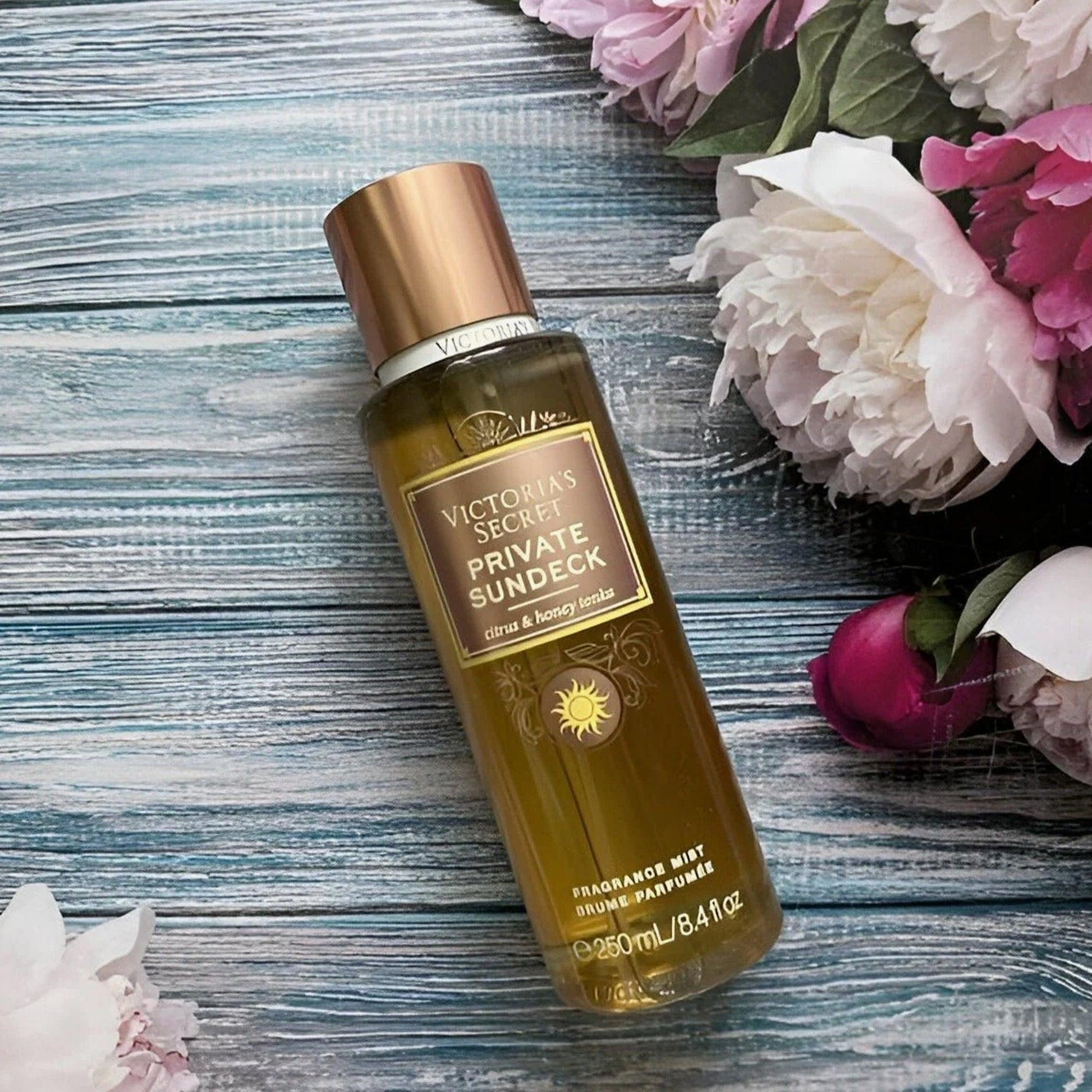 Victoria’s Secret Private Sundeck fragrance mist 250ml bottle placed on a wooden surface surrounded by pink and white peony flowers