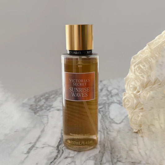Victorias Secret Sunrise Waves fragrance mist 250ml bottle with golden cap placed on a marble surface beside white rose flowers