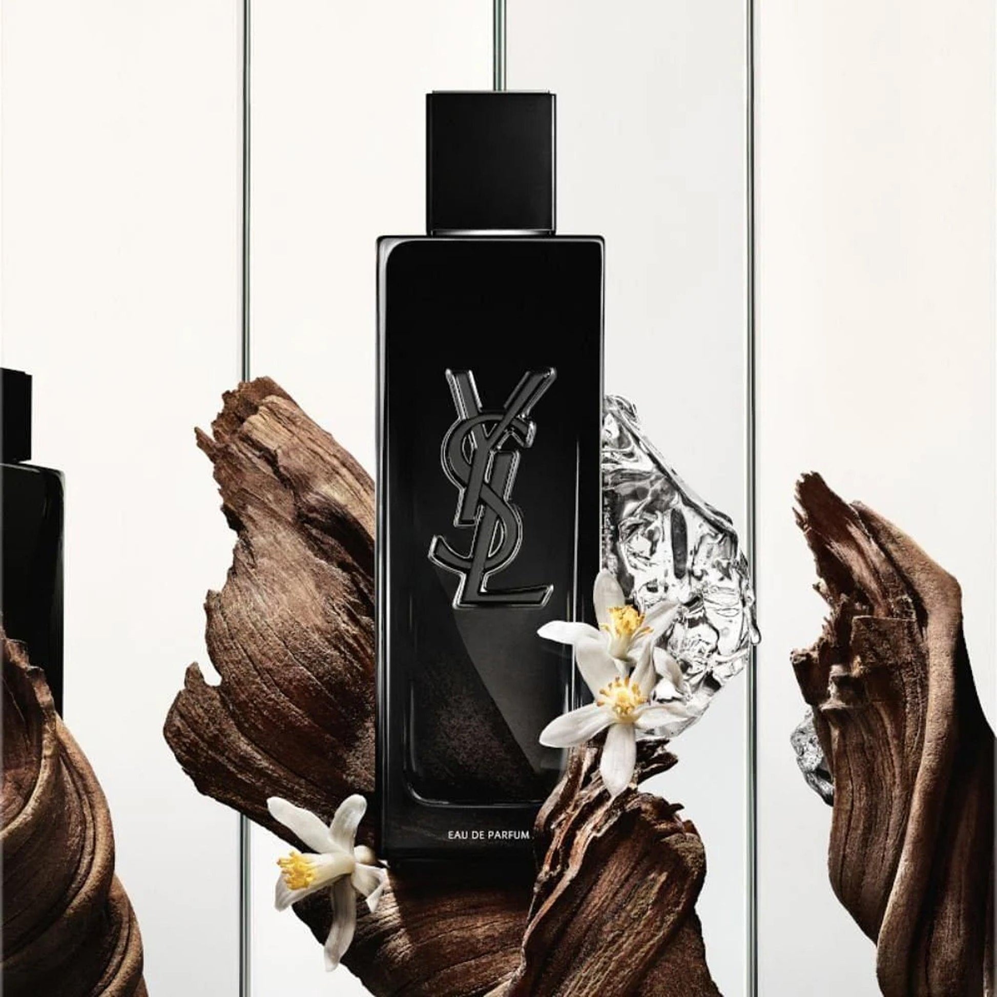 Yves Saint Laurent Y Eau De Parfum black bottle with YSL logo displayed among wood textures, ice, and white floral accents on a bright background